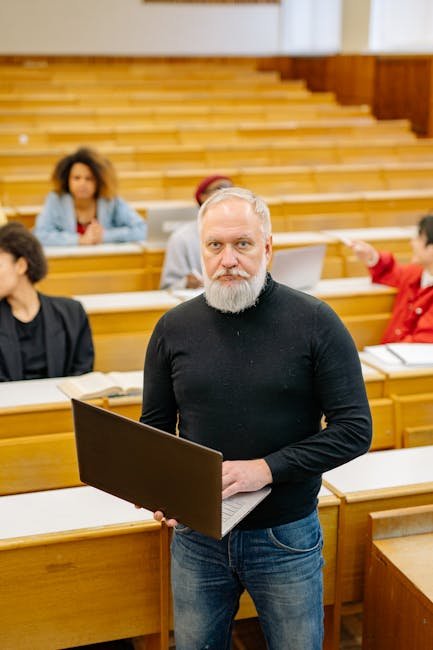 Senior male professor in a classroom holding a laptop, educating university students.